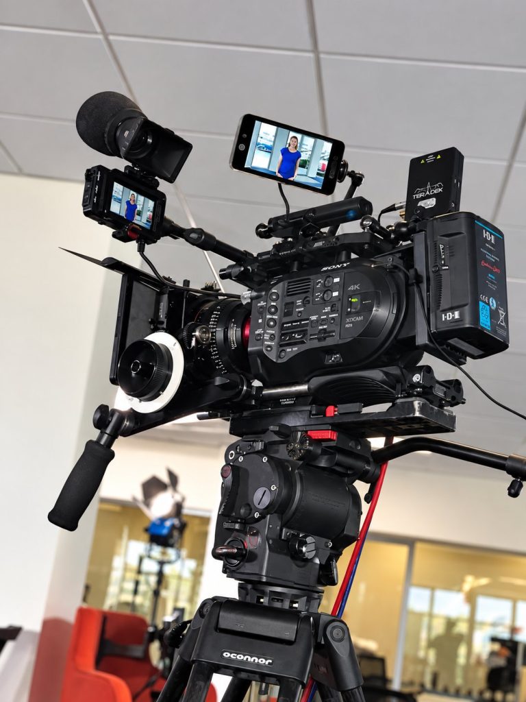 A broadcast camera with a presenter shown on its monitors
