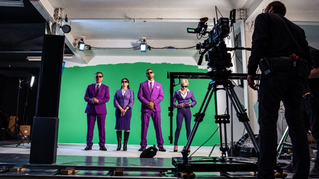 Four actors in purple suits and sunglasses being filmed against a green screen